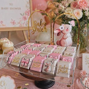 Könnte beinhalten: Eine Babyparty-Dekoration mit rosa und goldenen Akzenten. Die Szene umfasst ein Willkommensschild, Luftballons, Cupcakes und eine transparente Schale mit Pralinen, die mit rosa Schleifen und Hasenillustrationen verziert sind. Der Text "Baby Smier" ist zu sehen.
