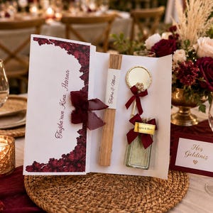 Pode incluir: Um conjunto de presente em cartolina branca com borda floral bordô, um leque de madeira, um pequeno espelho redondo com borda dourada e um frasco de vidro retangular. O conjunto é amarrado com uma fita bordô e inclui o texto Tuğba'nın Kına Hatırası e Tuğba & Mert.