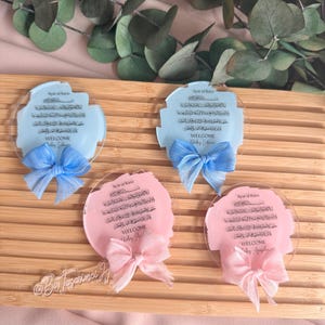May include: Four round acrylic plaques with a light blue or pink background. Each plaque features Arabic calligraphy and the text "Ayat al Kursi" and "Welcome". The plaques are decorated with a blue or pink ribbon bow.