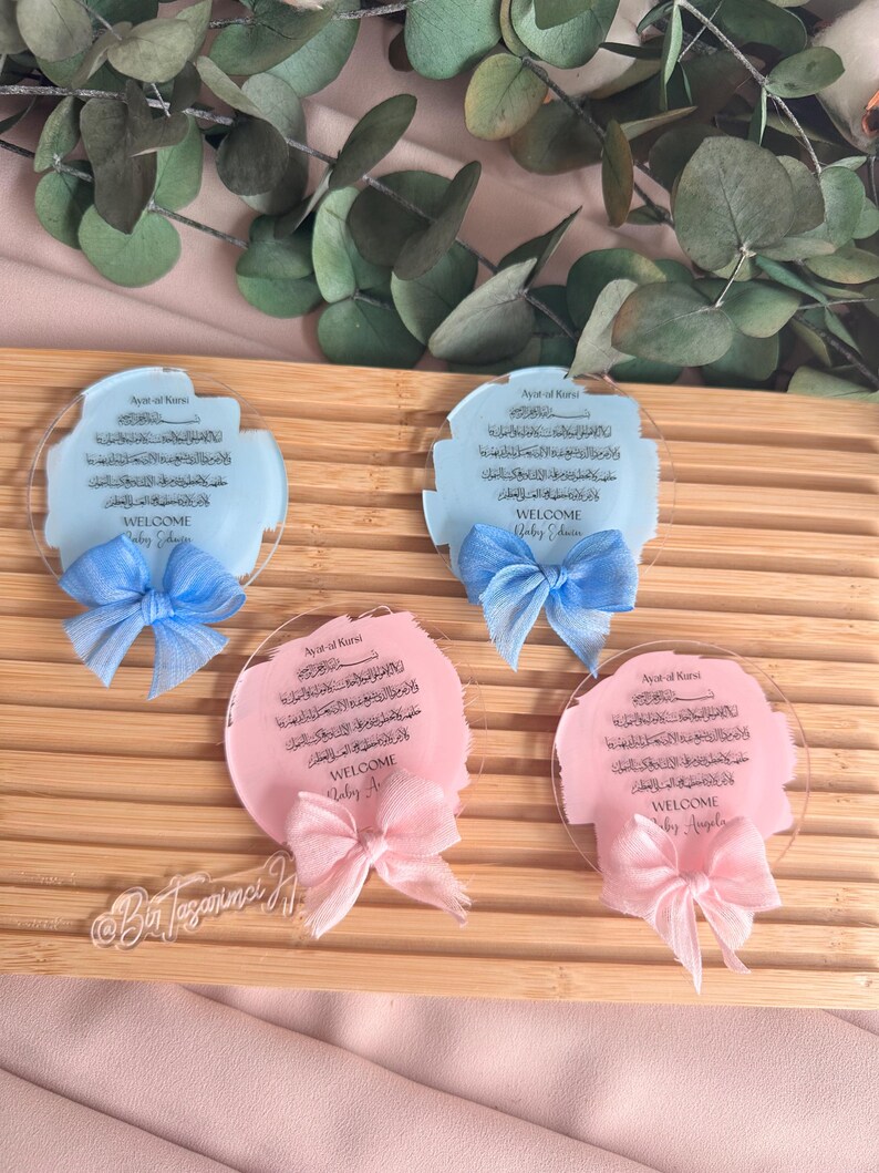 May include: Four round acrylic plaques with a light blue or pink background. Each plaque features Arabic calligraphy and the text "Ayat al Kursi" and "Welcome". The plaques are decorated with a blue or pink ribbon bow.