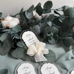 May include: Three clear acrylic wedding favor tags with white ribbon bows and pearl accents. The tags feature the text "Singe & Ahmet 01.02.2025 Nikah Hatira" or "Nikah Hat".