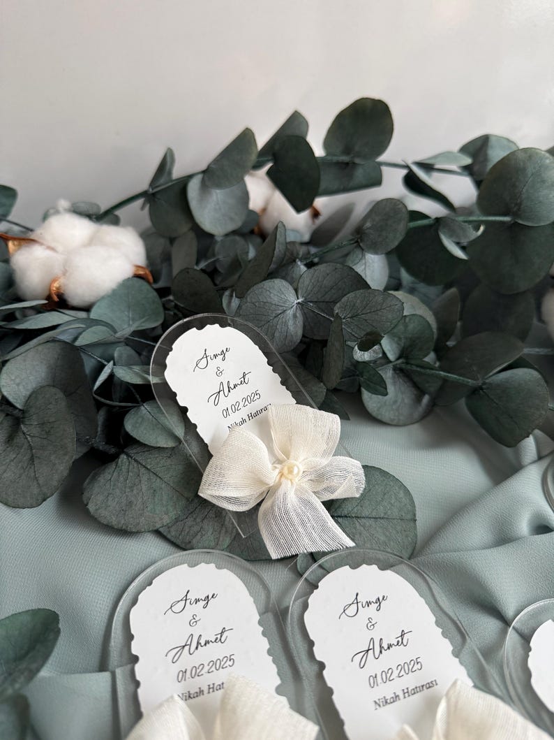 May include: Three clear acrylic wedding favor tags with white ribbon bows and pearl accents. The tags feature the text "Singe & Ahmet 01.02.2025 Nikah Hatira" or "Nikah Hat".
