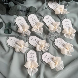 May include: A set of ten clear acrylic wedding favor tags with white satin ribbon bows and pearl accents. Each tag features the text "Singe & About" with a wedding date.