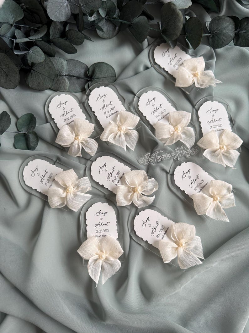 May include: A set of ten clear acrylic wedding favor tags with white satin ribbon bows and pearl accents. Each tag features the text "Singe & About" with a wedding date.
