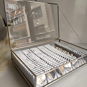 May include: A silver glass box with a hinged lid and a silver chain. The lid has a mirrored surface with the names "Rayda & Furkan" written in cursive. The box is filled with small silver heart-shaped favours, each with the names "Rayda & Furkan" and a date.