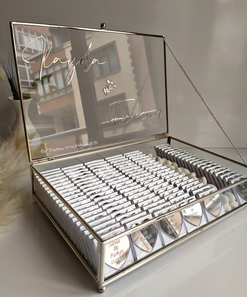 May include: A silver glass box with a hinged lid and a silver chain. The lid has a mirrored surface with the names "Rayda & Furkan" written in cursive. The box is filled with small silver heart-shaped favours, each with the names "Rayda & Furkan" and a date.