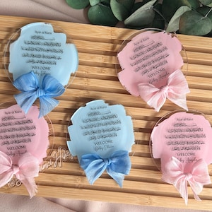 May include: Four clear acrylic plaques with a light blue or pink background and a black calligraphy design. Each plaque has a white ribbon tied around it and the text "Ayat-al Kursi" and "Welcome" with a baby's name.