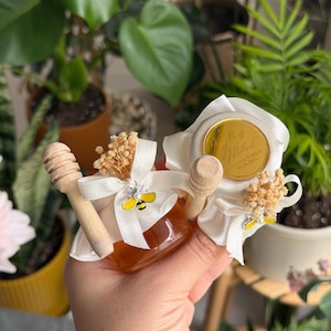 Könnte beinhalten: Kleines Glas Honig mit einem hölzernen Honiglöffel, gebunden mit einem weißen Band und einem gelben Bienenanhänger. Das Glas wird vor einem Hintergrund aus grünen Pflanzen gehalten.
