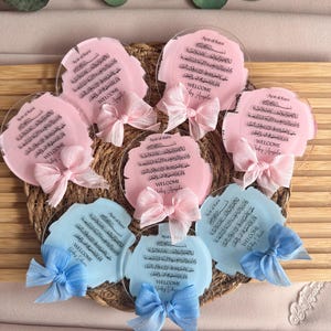 May include: A set of six round, clear acrylic plaques with pink and blue watercolor backgrounds. Each plaque features Arabic calligraphy and the text "Welcome Baby Angela" or "Welcome Baby Eduan". The plaques are decorated with pink and blue bows.