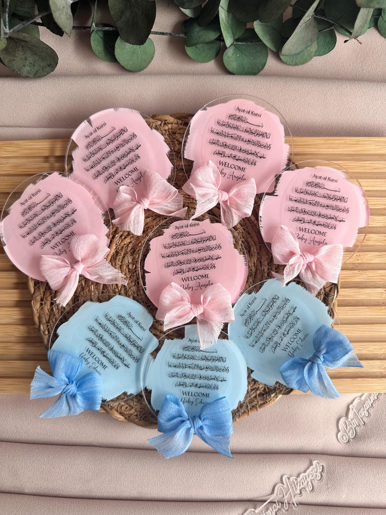 May include: A set of six round, clear acrylic plaques with pink and blue watercolor backgrounds. Each plaque features Arabic calligraphy and the text "Welcome Baby Angela" or "Welcome Baby Eduan". The plaques are decorated with pink and blue bows.