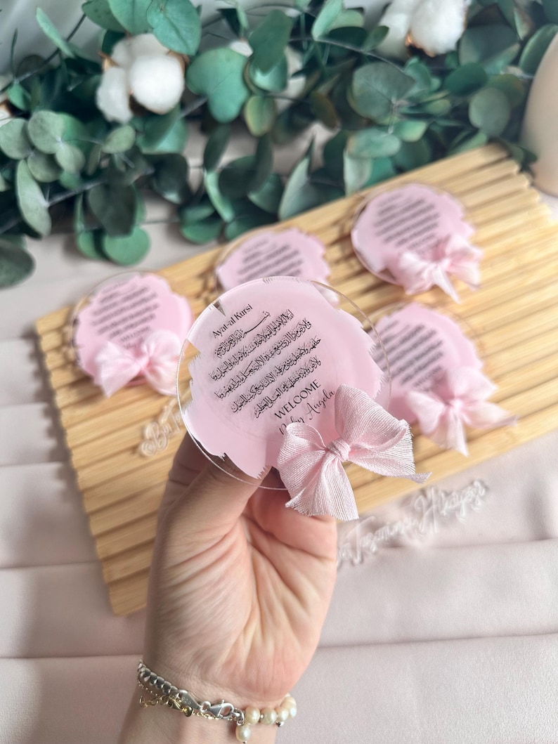 May include: Pink and white welcome sign with Arabic calligraphy and the text "Ayat-al Kursi" and "WELCOME Baby Angela".
