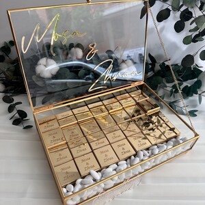 May include: A glass box with a gold trim and a gold chain. The box is filled with small gold boxes that have the names "Mosa & Zhraa" engraved on them. The box is surrounded by white stones.
