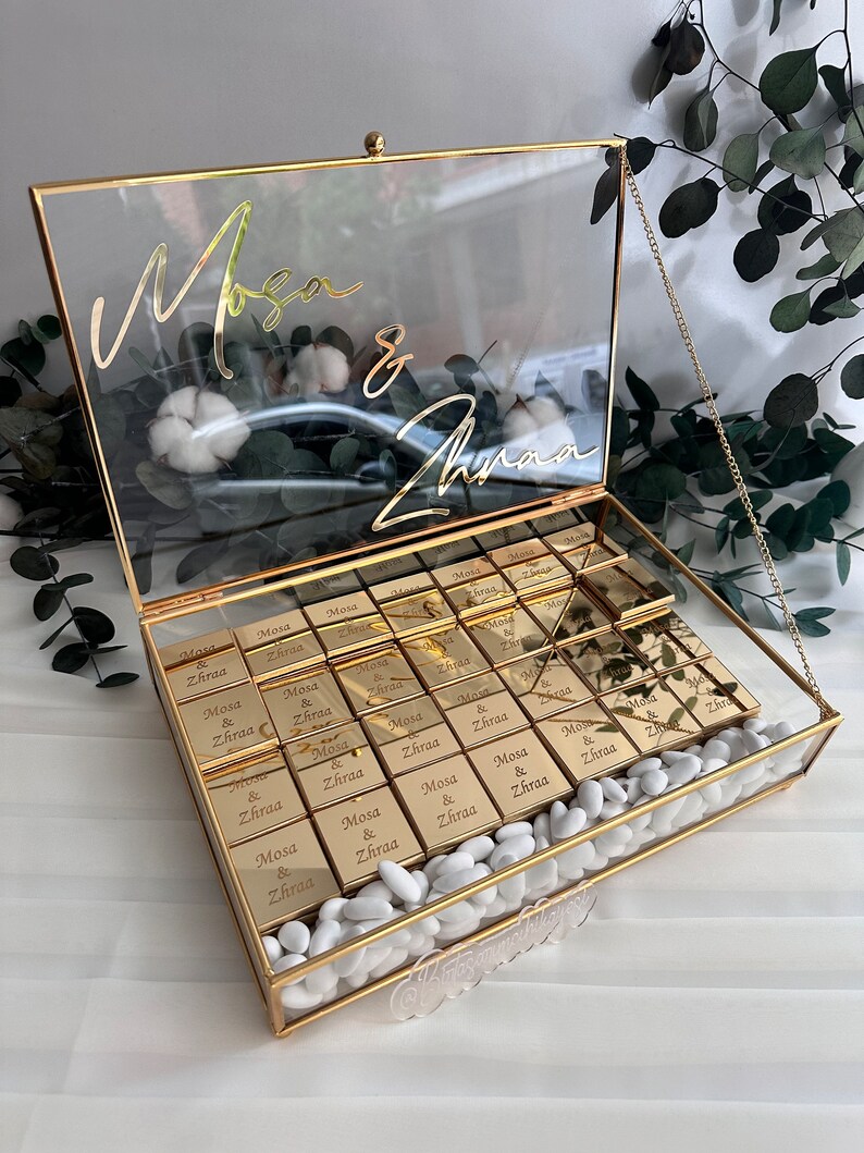 May include: A glass box with a gold trim and a gold chain. The box is filled with small gold boxes that have the names "Mosa & Zhraa" engraved on them. The box is surrounded by white stones.