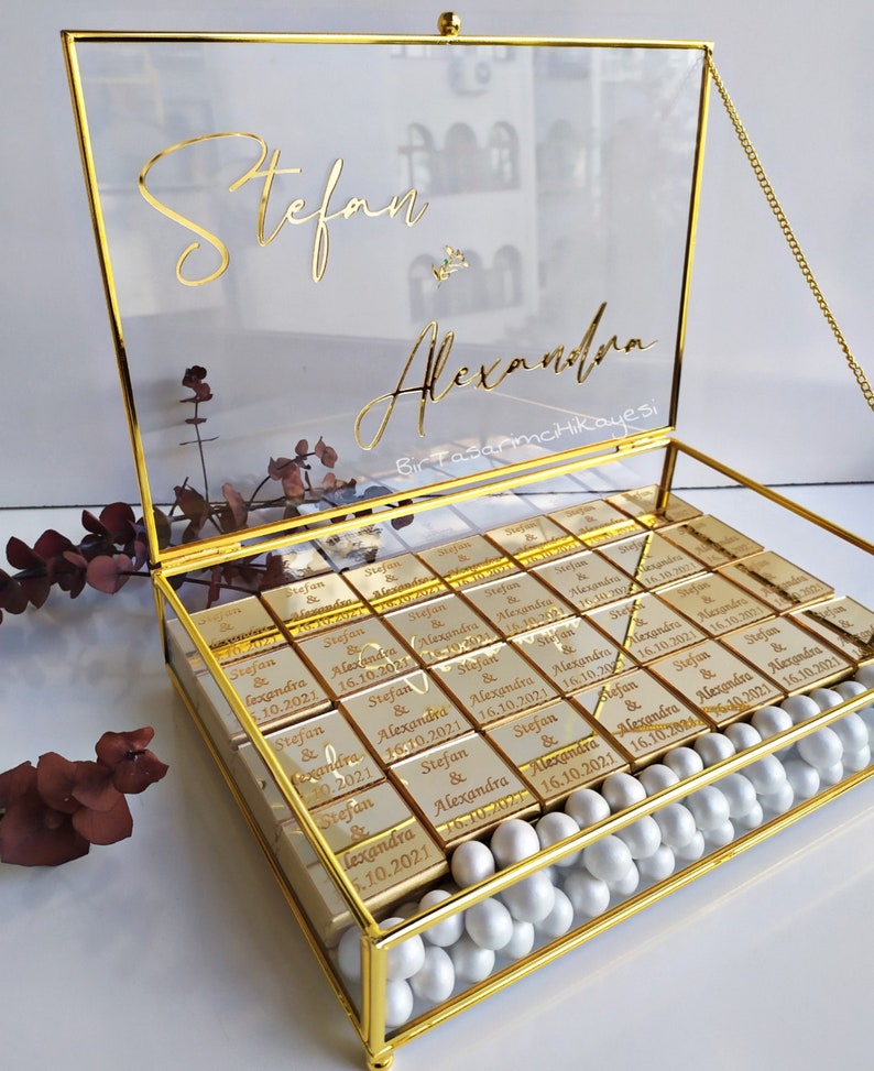 May include: A gold glass box with a lid filled with small gold-wrapped chocolates. Each chocolate has the names "Stefan & Alexandra" and the date "16.10.2021" printed on it. The lid of the box has the names "Stefan & Alexandra" written in gold cursive lettering.