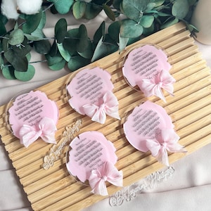 May include: Five pink and white circular acrylic plaques with Arabic calligraphy and the words "Welcome Baby Ayla" in black lettering. Each plaque has a pink ribbon tied around it.