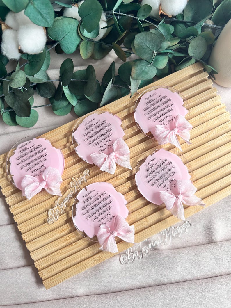 May include: Five pink and white circular acrylic plaques with Arabic calligraphy and the words "Welcome Baby Ayla" in black lettering. Each plaque has a pink ribbon tied around it.