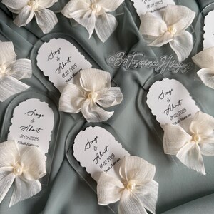 May include: Clear acrylic wedding favor with a white bow and pearl. The favor has the text "Singe & Ahmet 01.02.2025 Nikah Hatırası" printed on it.