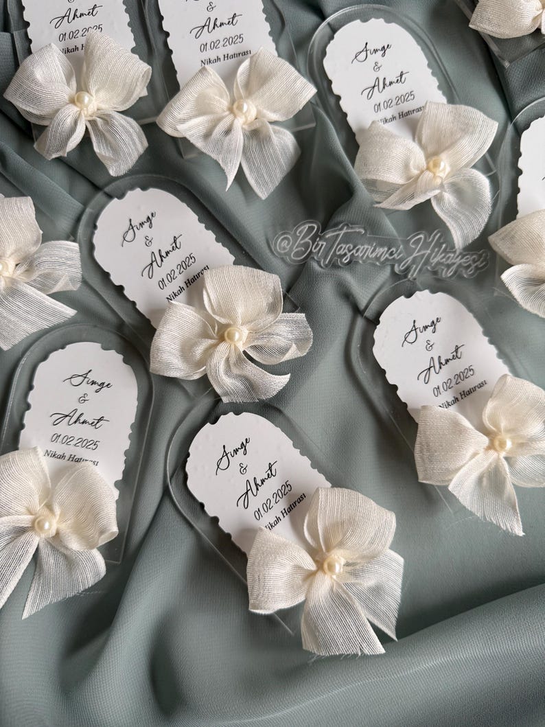 May include: Clear acrylic wedding favor with a white bow and pearl. The favor has the text "Singe & Ahmet 01.02.2025 Nikah Hatırası" printed on it.