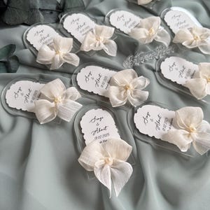 May include: A set of clear acrylic wedding favors with white ribbon bows and pearl accents. Each favor features the names "Singe & Ahmet" and the wedding date "01.02.2025".