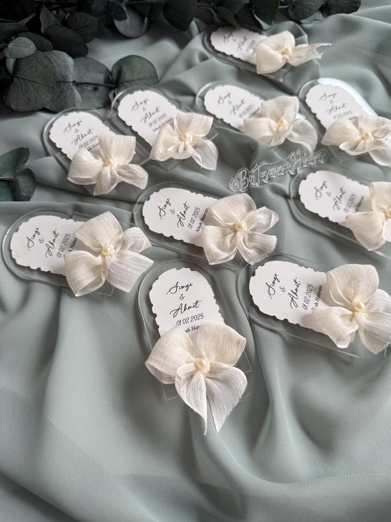 May include: A set of clear acrylic wedding favors with white ribbon bows and pearl accents. Each favor features the names "Singe & Ahmet" and the wedding date "01.02.2025".