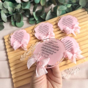 May include: Pink and white welcome sign with Arabic calligraphy and the text "Welcome by Angel" on a clear acrylic base. The sign is decorated with a pink ribbon.