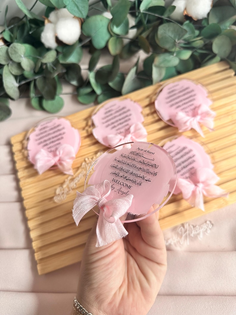 May include: Pink and white welcome sign with Arabic calligraphy and the text "Welcome by Angel" on a clear acrylic base. The sign is decorated with a pink ribbon.
