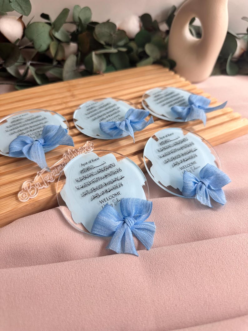 May include: Five clear acrylic plaques with blue ribbons and Arabic calligraphy. The plaques feature the text "Ayat al Kursi" and "Welcome Baby Edition".