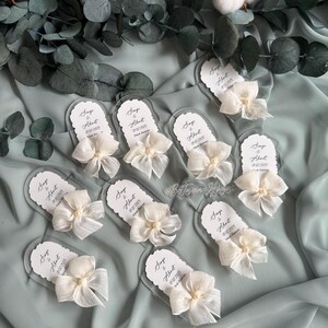 May include: A set of nine clear acrylic wedding favor tags with white satin bows and pearl accents. Each tag features the text "Singe & About" with a wedding date.