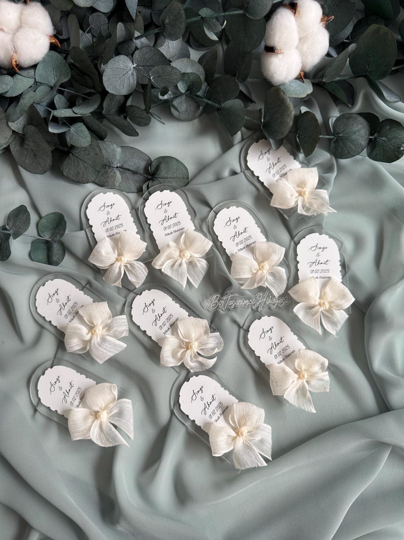 May include: A set of nine clear acrylic wedding favor tags with white satin bows and pearl accents. Each tag features the text "Singe & About" with a wedding date.