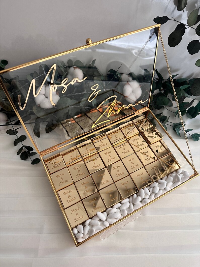 May include: A gold glass box with a gold chain handle filled with small gold boxes that say "Mosa & Zhraa" and white candy. The lid of the box has the names "Mosa & Zhraa" written in gold cursive.