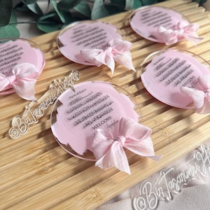 May include: Pink and white acrylic round ornaments with Arabic calligraphy and a pink ribbon bow. The text reads "Ayat-al Kursi" and "Welcome Angela".