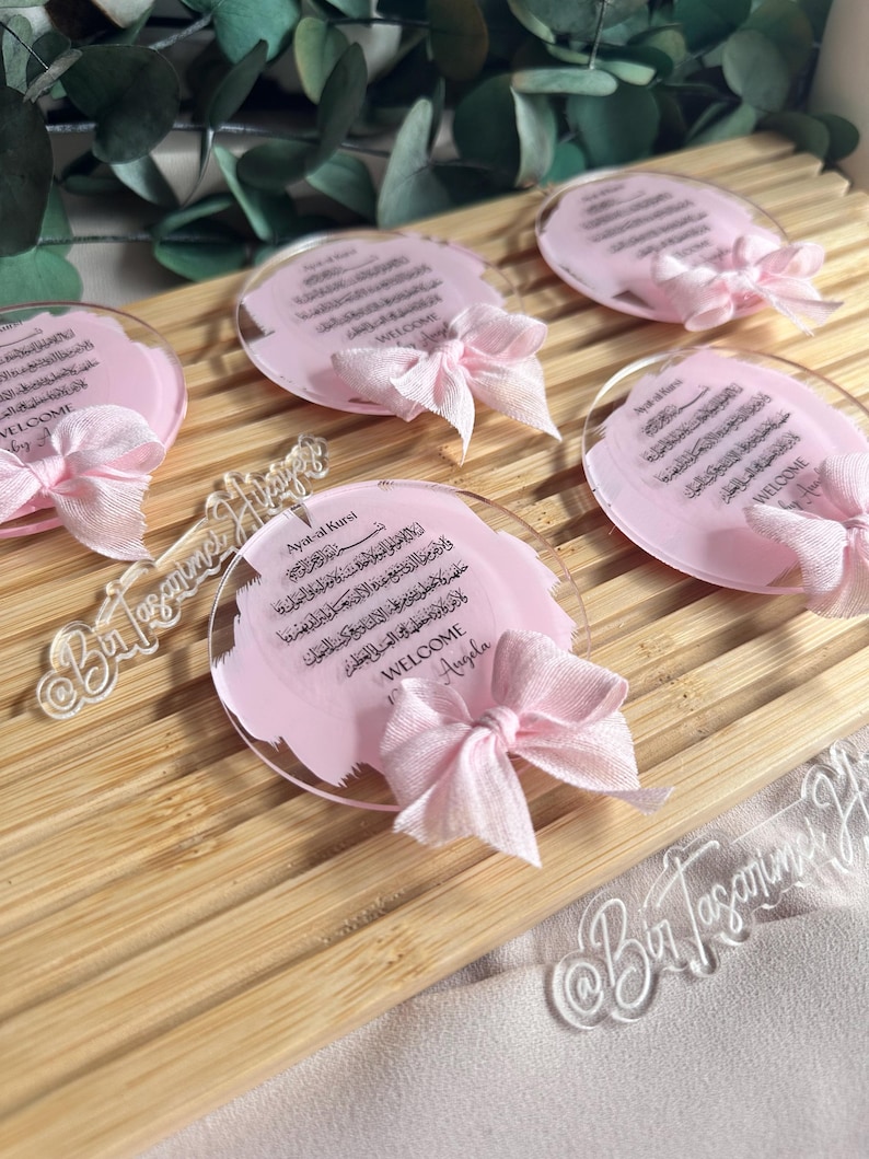May include: Pink and white acrylic round ornaments with Arabic calligraphy and a pink ribbon bow. The text reads "Ayat-al Kursi" and "Welcome Angela".