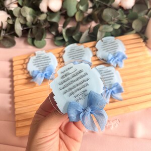 May include: A set of blue and white baby shower favors with a clear acrylic backing. The favors feature a blue ribbon and the Arabic text "Ayat-al Kursi" and "WELCOME Baby Edu".