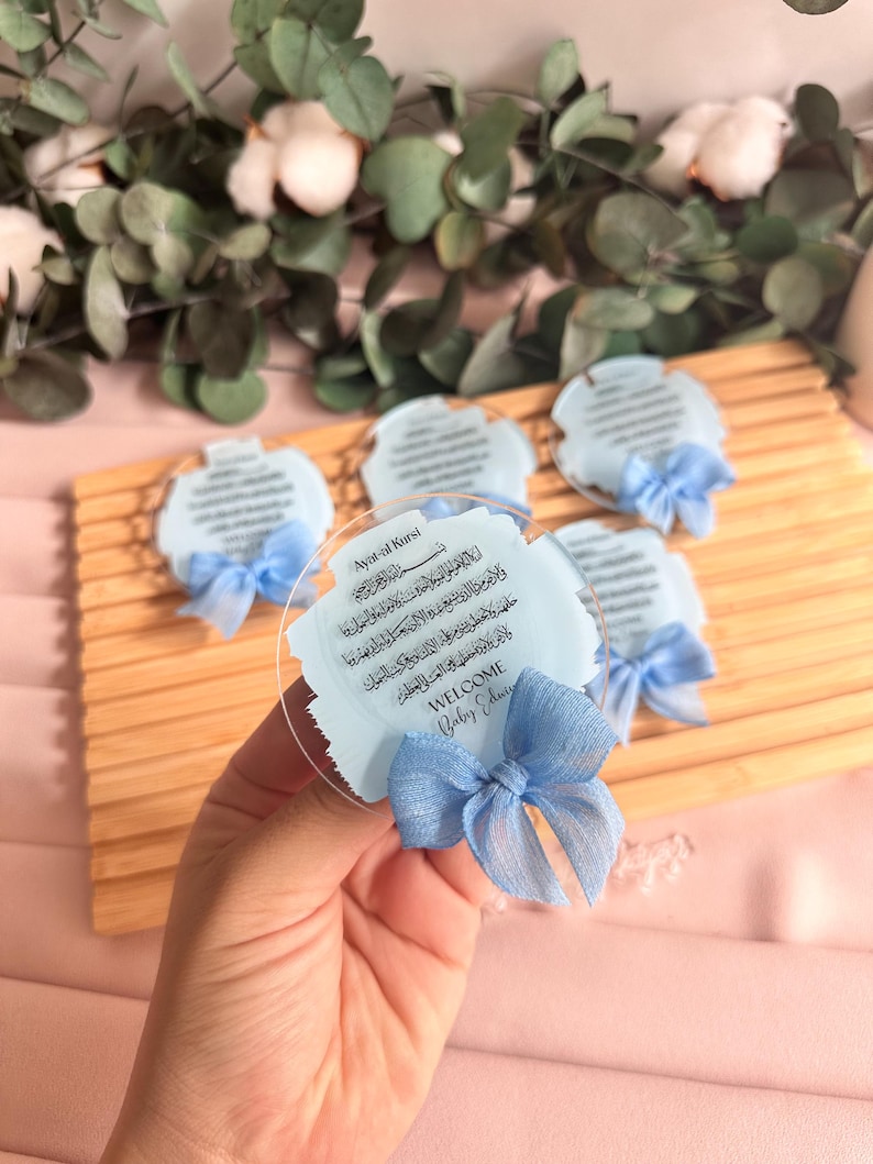 May include: A set of blue and white baby shower favors with a clear acrylic backing. The favors feature a blue ribbon and the Arabic text "Ayat-al Kursi" and "WELCOME Baby Edu".