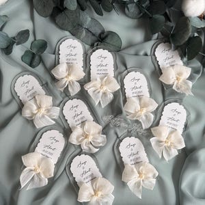 May include: A set of nine clear acrylic wedding favors with white bows and pearls. Each favor has a white card with the text "Singe & About 01.02.2025 Nikah Mat" printed on it.