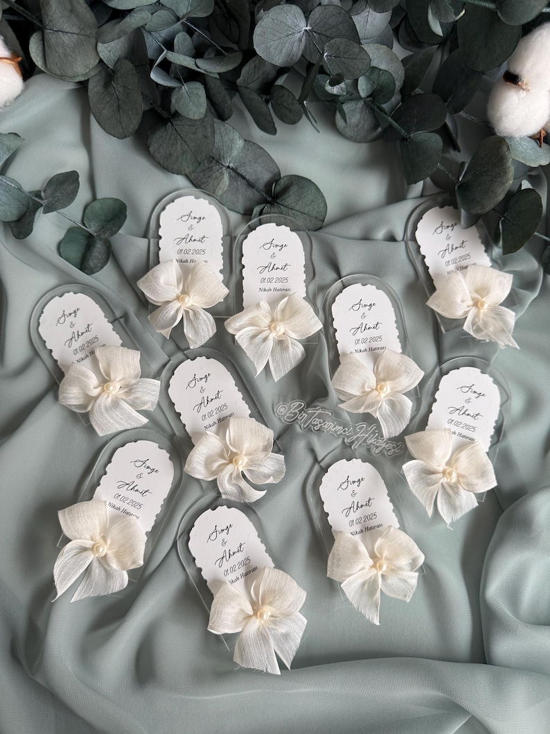 May include: A set of nine clear acrylic wedding favors with white bows and pearls. Each favor has a white card with the text "Singe & About 01.02.2025 Nikah Mat" printed on it.