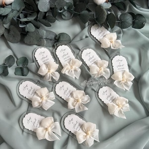 May include: A set of ten clear acrylic wedding favor tags with white satin bows and pearl accents. Each tag features the names "Sage & Albert" and the wedding date "07.02.2025".
