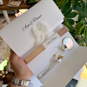May include: A white invitation set with the names "Aya & Timur" printed in black script. The set includes a fan, a small mirror, and a perfume bottle, all adorned with ribbons. The fan is made of light brown wood.