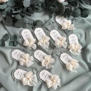 May include: A collection of clear acrylic wedding favor tags with white satin bows and pearl accents. Each tag features the names "Sage & Albert" and the wedding date "02.02.2025".