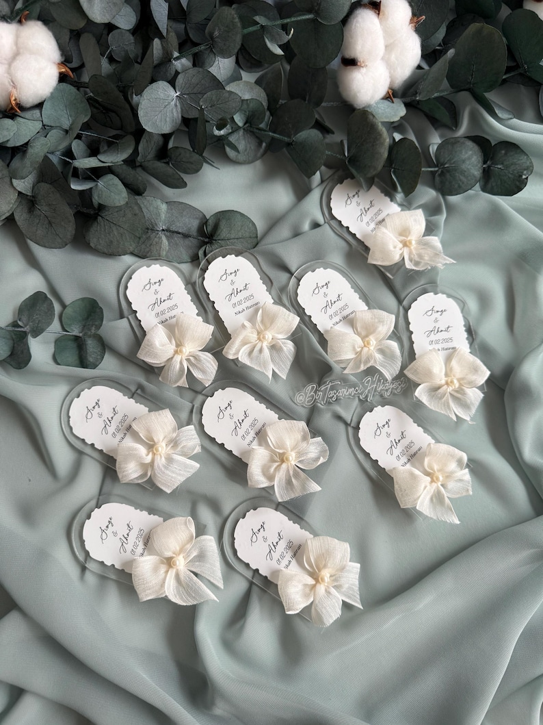 May include: A collection of clear acrylic wedding favor tags with white satin bows and pearl accents. Each tag features the names "Sage & Albert" and the wedding date "02.02.2025".