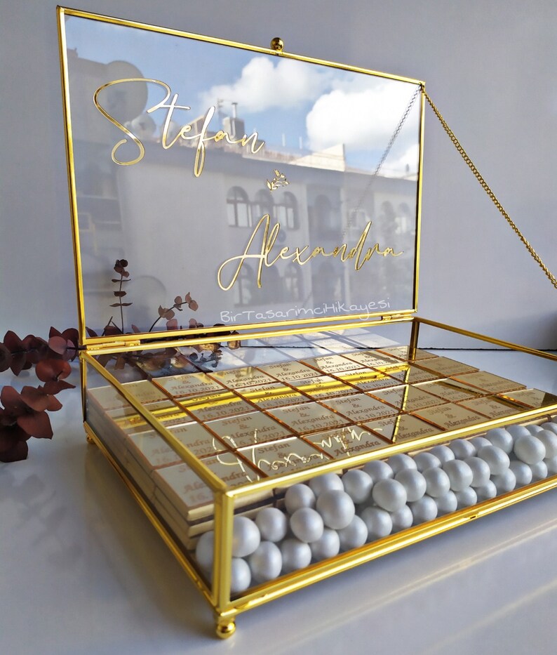 May include: A gold glass box with a hinged lid and a chain. The lid has the names "Stefan & Alexander" written in gold script. The box is filled with small gold-wrapped chocolates and white pearl-like sweets.