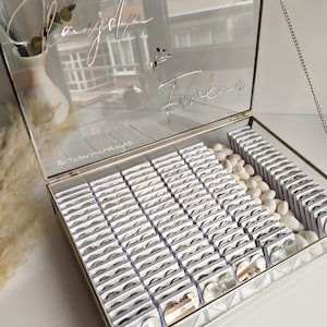 May include: A glass box filled with small silver heart-shaped favours with the text "İlayda & Furkan 23.12.2023" on them. The box is decorated with a silver chain and the names "İlayda & Furkan" are written on the lid.