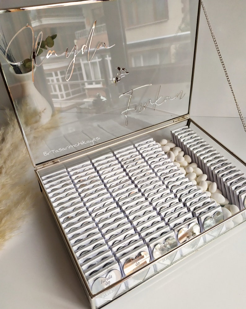 May include: A glass box filled with small silver heart-shaped favours with the text "İlayda & Furkan 23.12.2023" on them. The box is decorated with a silver chain and the names "İlayda & Furkan" are written on the lid.