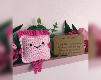 May include: Pink crocheted worry monster toy with a smiling face and small legs. The toy is sitting on a shelf with a brown card that reads "Worry Monster I'm your little worry monster. I'll be here night and day. Tell me all your worries and I'll eat them all away!"
