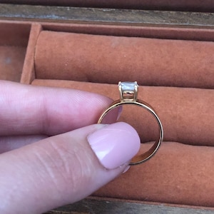 May include: A gold band engagement ring with a single, clear, square-shaped diamond.