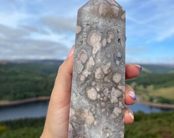 May include: A clear, faceted crystal point with a unique, swirling pattern of white and pink inclusions. The crystal is held in a hand against a backdrop of a blue sky and green hills.