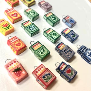 May include: A selection of small matchbox charms in diverse colours and patterns. Each charm displays a unique design, such as hearts, flowers, and strawberries. Some matchboxes have text that reads "FUCK THIS" and "LET'S GO".