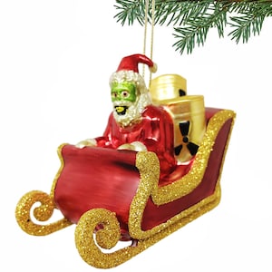 May include: A red glass Christmas ornament of a zombie Santa Claus in a sleigh with a green face and a white beard. He is holding a red bag and there are two yellow barrels with a radiation symbol on them in the sleigh.