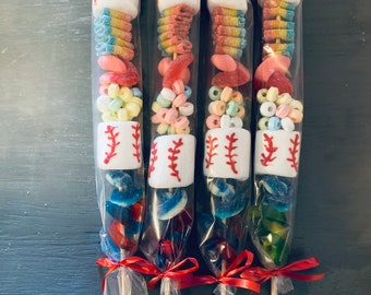 Pode incluir: Quatro sacos de plástico transparente cheios de doces coloridos e coroados com um marshmallow branco em forma de bola de beisebol com costuras vermelhas. Os sacos são amarrados com uma fita vermelha e presos a palitos de madeira.