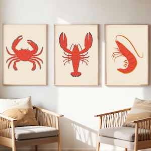 Op de afbeelding: Drie ingelijste kunstprints met illustraties van zeevruchten. De prints tonen een rode krab, een rode kreeft en een rode garnaal op een crèmekleurige achtergrond. De frames zijn licht van hout en de kunst wordt op een witte muur getoond.